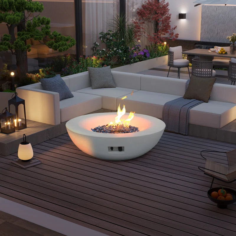 Outdoor concrete natural gas fire pit bowl on a modern patio with wooden decking, surrounded by white sectional seating and garden decor.